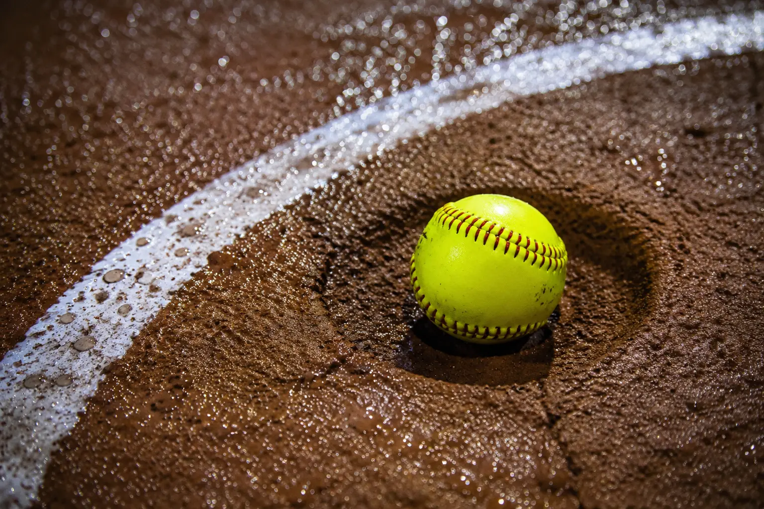 Yellow softball resting in damp infield clay beside a chalk arc